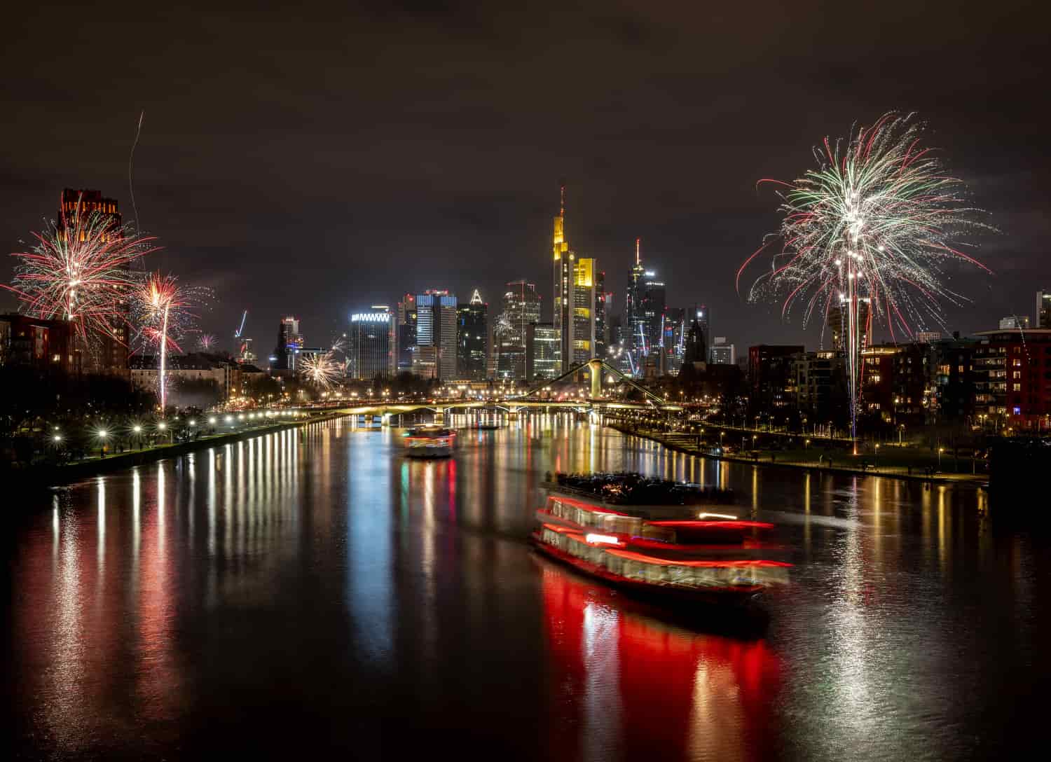 Los barcos de fiesta cruzan el río Main con solo unos pocos fuegos artificiales cerca de los edificios del distrito bancario en Frankfurt, Alemania, la madrugada del sábado 1 de enero de 2022. Debido a la pandemia de coronavirus, no se permitieron los fuegos artificiales.