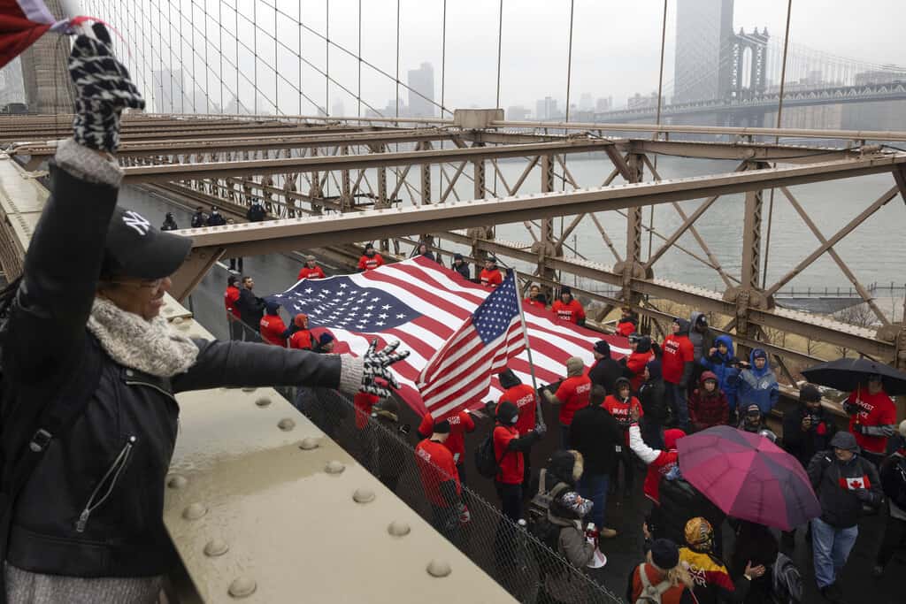 Personas con banderas estadounidenses marchan en el puente de Brooklyn durante una protesta contra el mandato de vacunas antes de la posible terminación de los empleados de la ciudad de Nueva York debido a su estado de vacunación, el lunes 7 de febrero de 2022, en Nueva York.