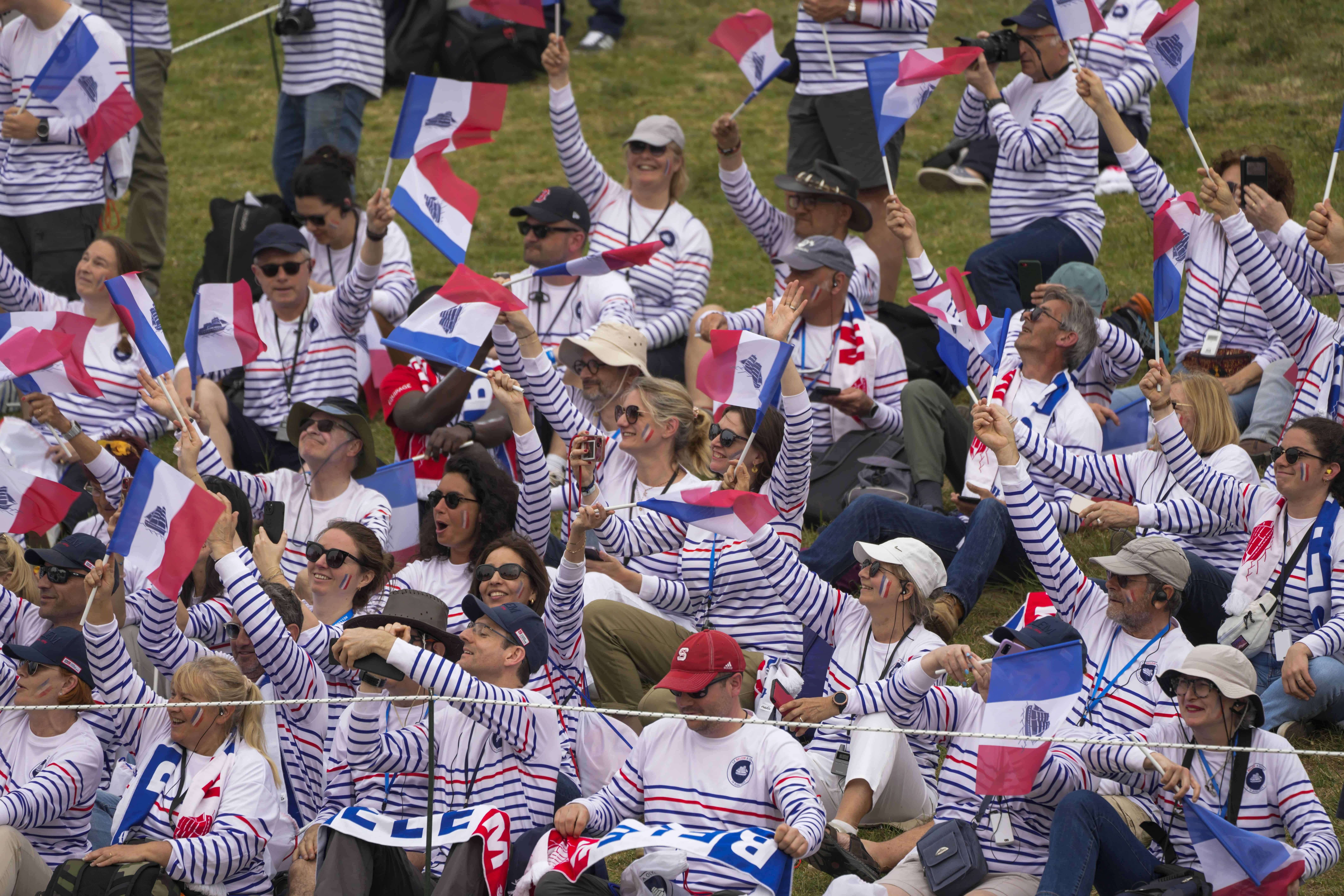 Los espectadores ondean banderas francesas con una imagen de la goleta francesa de tres mástiles Belem durante la ceremonia oficial de encendido de la llama de los Juegos Olímpicos de París, en el sitio de la Antigua Olimpia, Grecia, el martes 16 de abril de 2024.