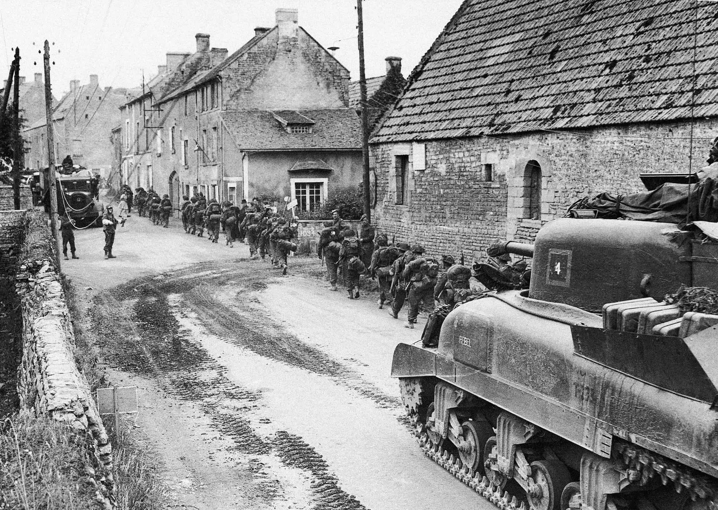 ARCHIVO - Tropas y vehículos británicos que pasan tierra adentro a través de una aldea en Normandía. Los linieros están ocupados colocando cables de comunicación mientras los blindados británicos, canadienses y estadounidenses los pasan en ruta hacia la línea del frente, el 13 de junio de 1944.