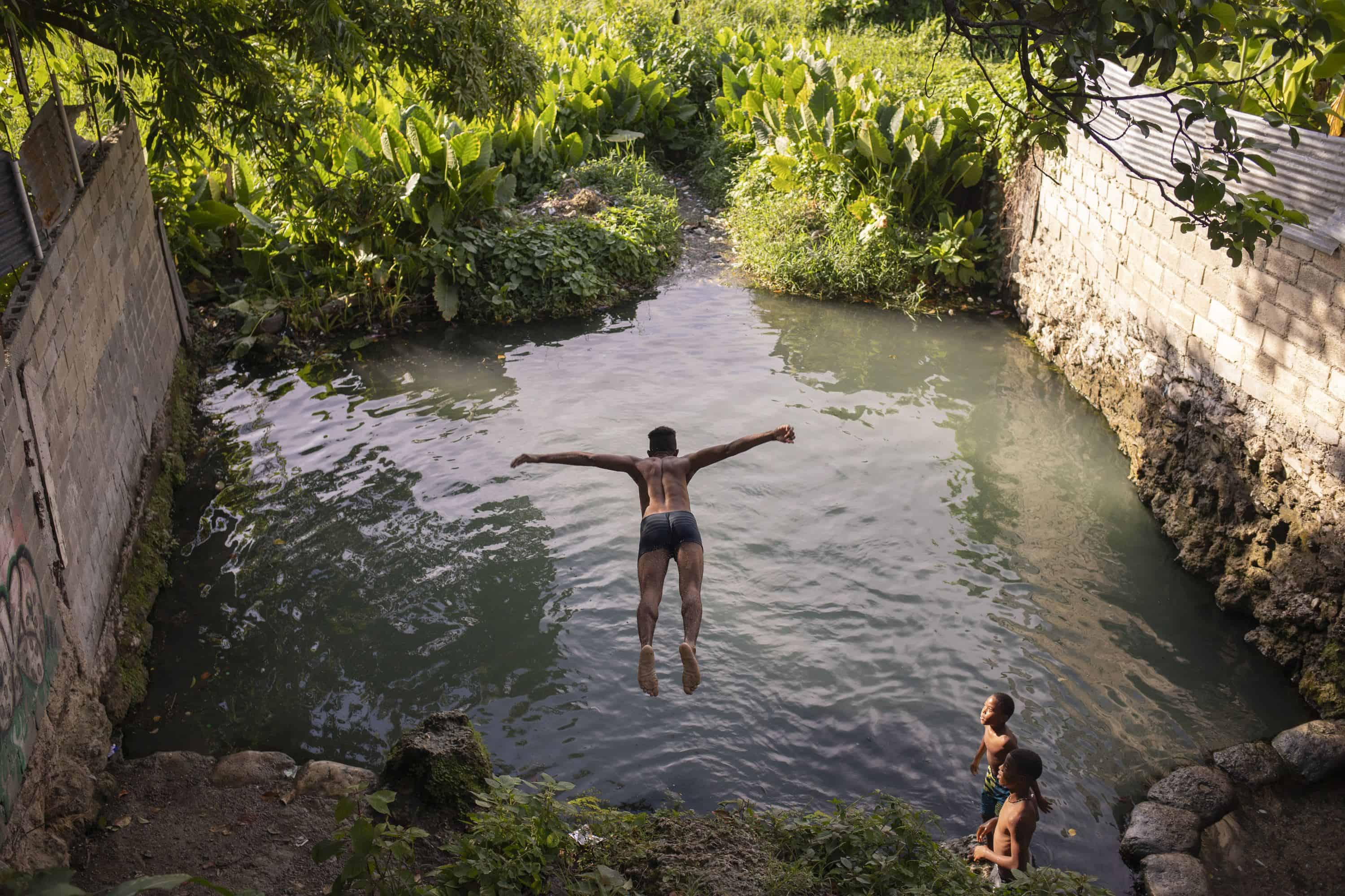 Categoría Vida Diaria: Primer lugar. Un grupo de jóvenes disfruta saltando y refrescándose en la poza de agua en el barrio Los Tres Brazos en Santo Domingo Este, República Dominicana.Esta imagen captura la alegría y la camaradería de la vida cotidiana en un barrio vibrante, mostrando cómo momentos simples pueden ser llenos de felicidad y comunidad.