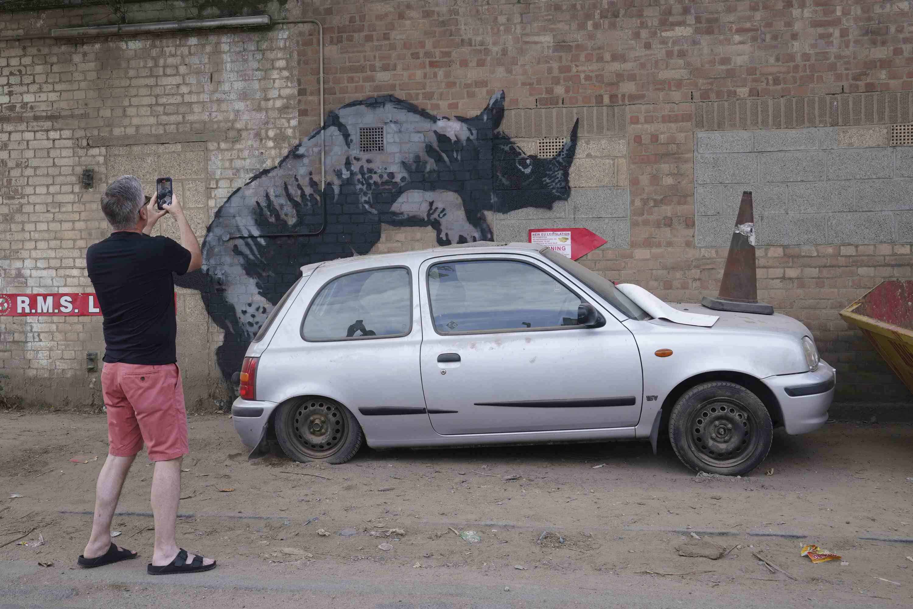 Un hombre fotografía una nueva obra de arte de Banksy, que representa un rinoceronte que parece subirse a un automóvil, la octava obra de arte de su colección de temática animal, en Westmoor Street en Charlton, sureste de Londres, el lunes 12 de agosto de 2024.