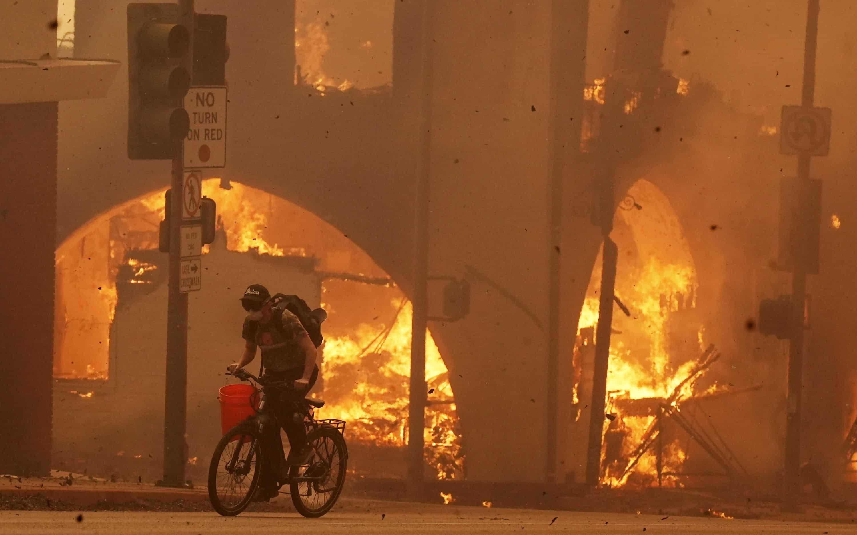 Un ciclista pedalea junto a una estructura en llamas en Lake Avenue, el miércoles 8 de enero de 2025, en la sección del centro de Altadena de Pasadena, California.