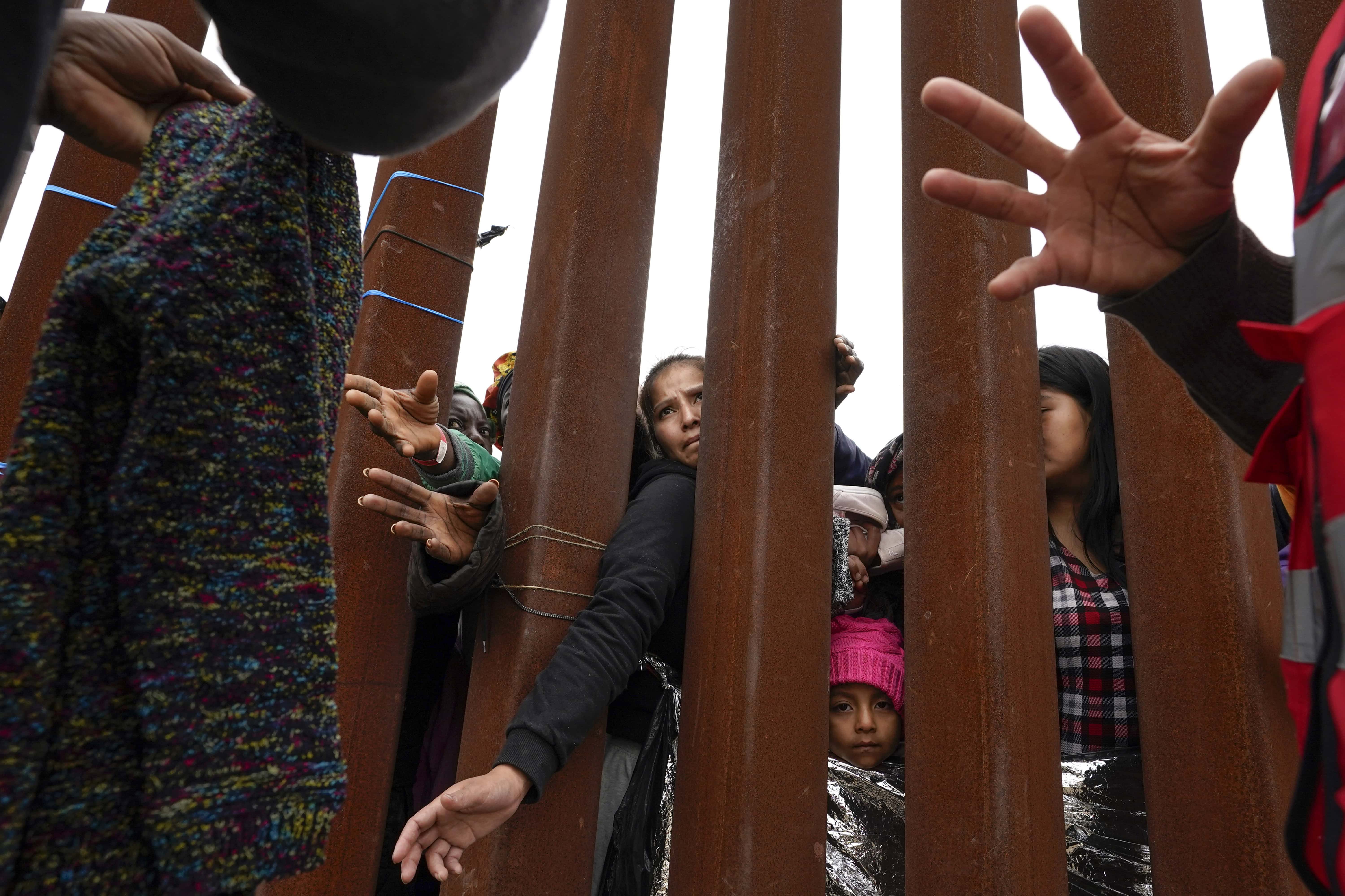 Migrantes extienden la mano a través del muro fronterizo que separa Estados Unidos de México para recibir las prendas que reparten los voluntarios entre solicitantes de asilo, el 12 de mayo de 2023, en San Diego. (AP