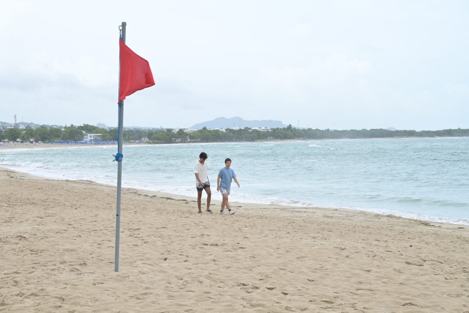 Una bandera roja en primer plano en una playa de Puerto Plata, mientras dos personas caminan bajo un cielo nublado. 