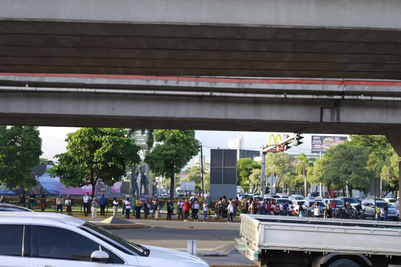 Personas hacen fila en espera de transporte en la avenida John F. Kennedy.