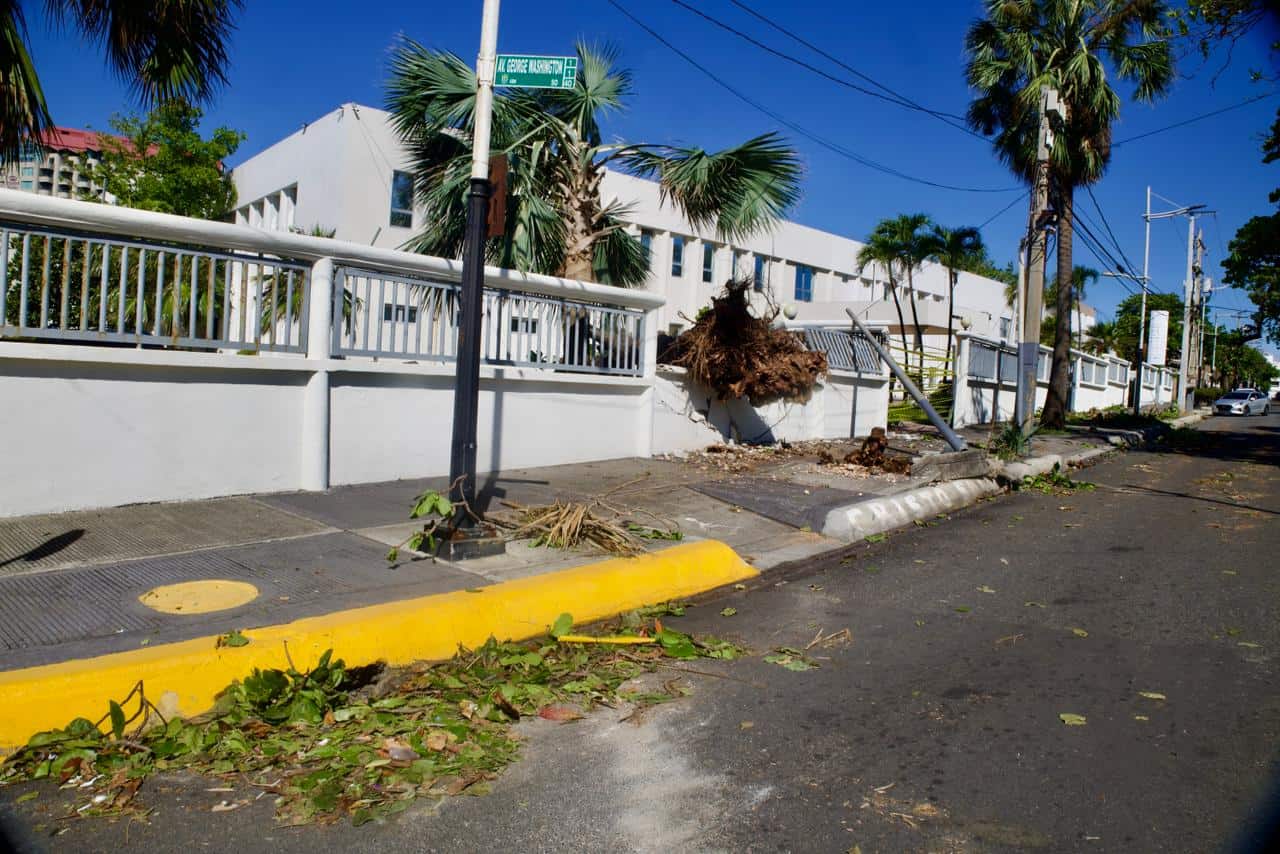 Restos de pared, cables expuestos y un poste afectado permanecen en la acera tras los daños causados por los fuertes vientos y lluvias.