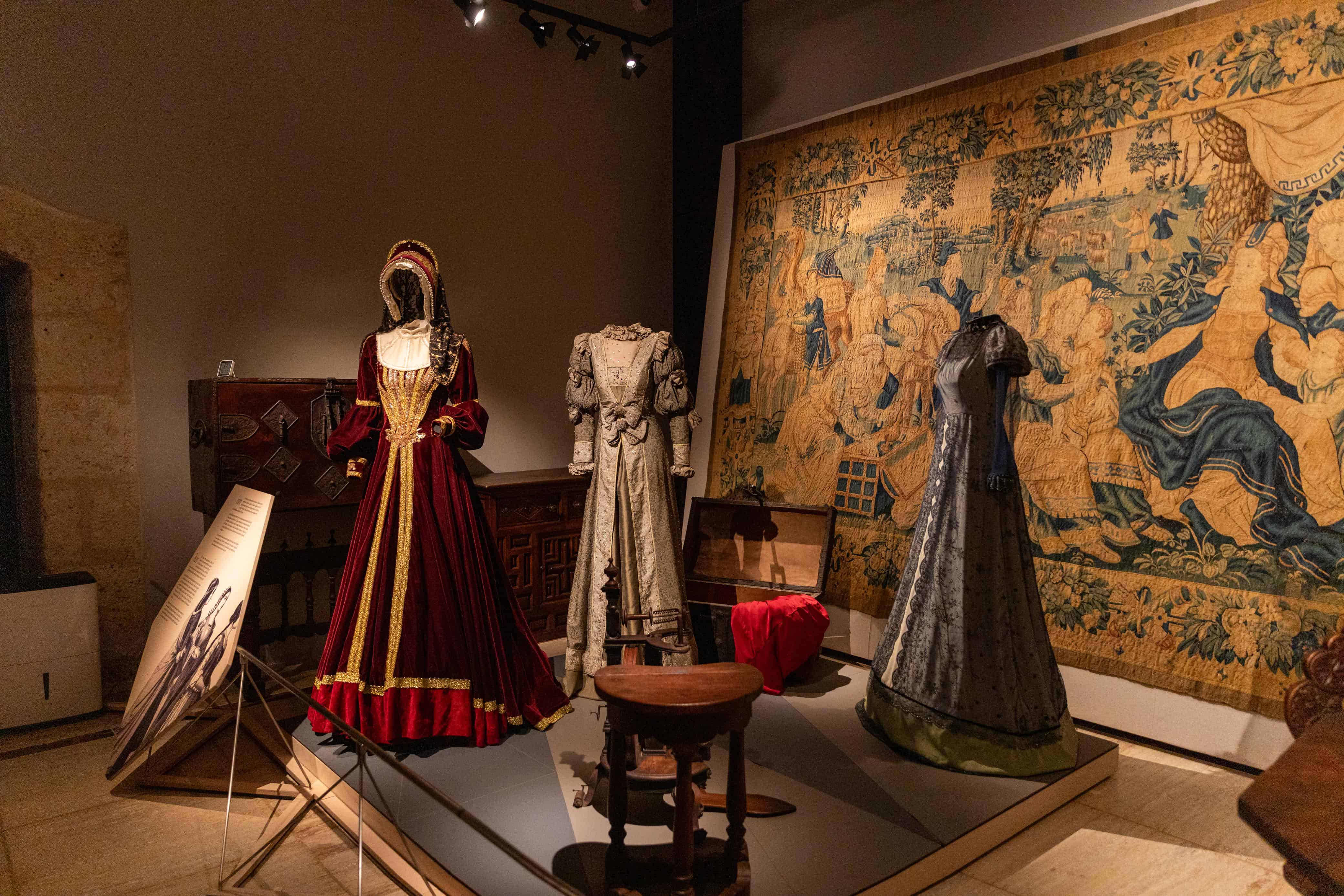 Sala de exhibici&oacute;n que muestra indumentaria hist&oacute;rica y trajes de &eacute;poca renacentista frente a un gran tapiz tejido con motivos florales y aleg&oacute;ricos.