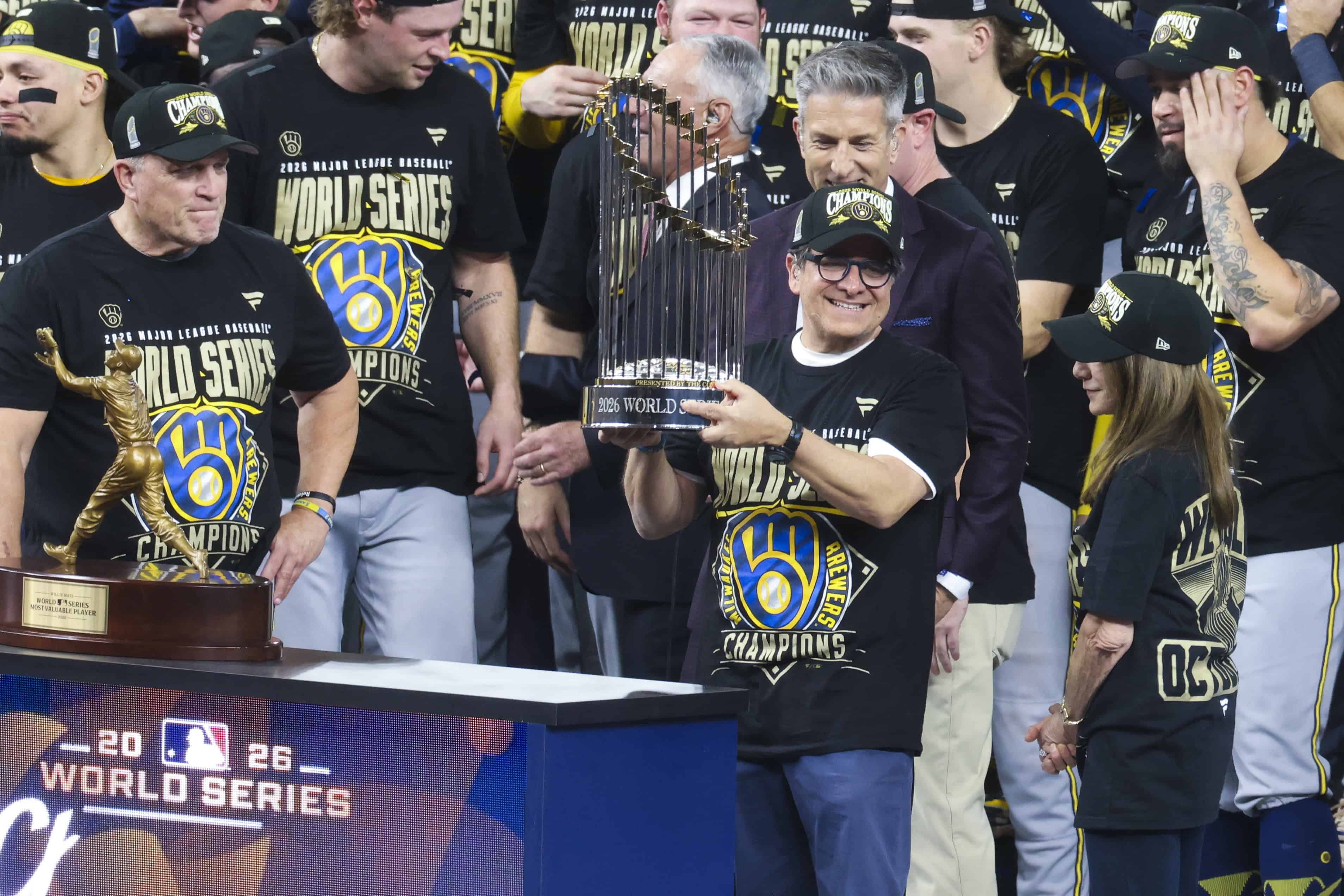El propietario de los Milwaukee Brewers, Mark Attanasio (C-D), sostiene el trofeo de campe&oacute;n tras la victoria en el quinto partido de la Serie Mundial de las Grandes Ligas de B&eacute;isbol (MLB) antelos Tampa Bay Rays en el Tropicana Field de San Petersburgo, Florida, EE. UU., el 28 de octubre de 2026. 