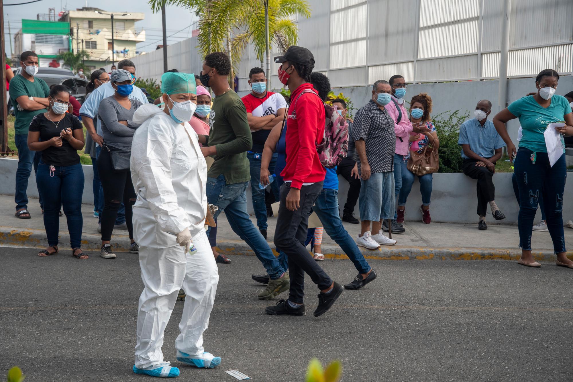 Un médico totalmente protegido, camina frente a un gr de personas que permanecen frente al hospital Moscoso Puello, esperando les sean tomadas las muestras de diagnóstico del coronavirus (PCR). Foto: Danelis Sena/Diario Libre.