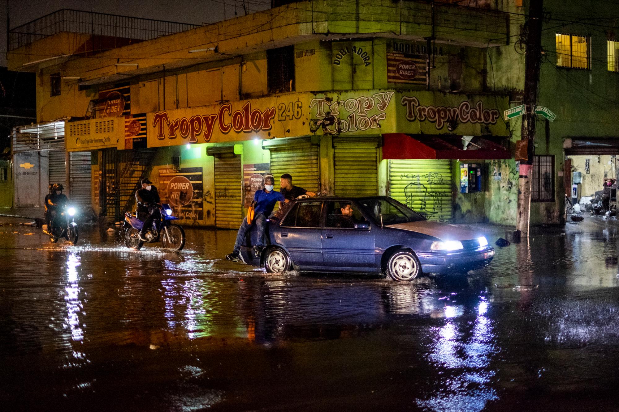 Santo Domingo, RD: dos hombres cruzan un charco de agua montados en la parte trasera de un auto en la Avenida Nicolás de Ovando esquina Juan Erazo en Villas Agrícolas