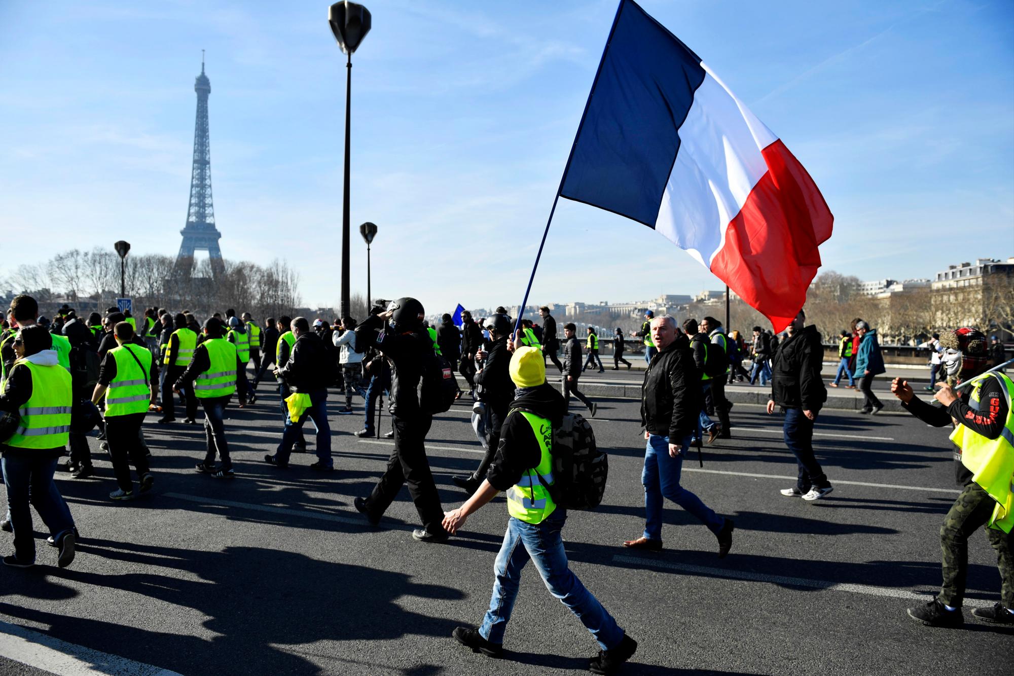 París (Francia), 16/02/2019.- Los manifestantes del movimiento ‘Gilets Jaunes’ (chalecos amarillos) que sostienen una bandera francesa pasan por la Torre Eiffel mientras caminan hacia los Inválidos durante la manifestación del ‘Acto XIV’ (la 14ª consecutiva protesta nacional un sábado) en París, Francia, el 16 de febrero de 2019. El llamado ‘Gilets Jaunes’ es un movimiento de protesta popular con partidarios de un amplio espectro del espectro político, que originalmente comenzó con una protesta en todo el país a fines de 2018. Contra los altos precios del combustible.