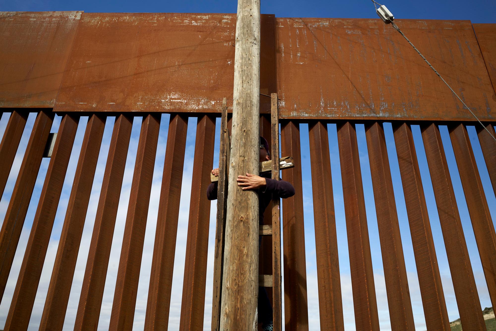 Un hombre utiliza una escalera para escalar un poste eléctrico en un vecindario fronterizo en Tijuana, México, junto a una sección recién reemplazada de la pared fronteriza. 