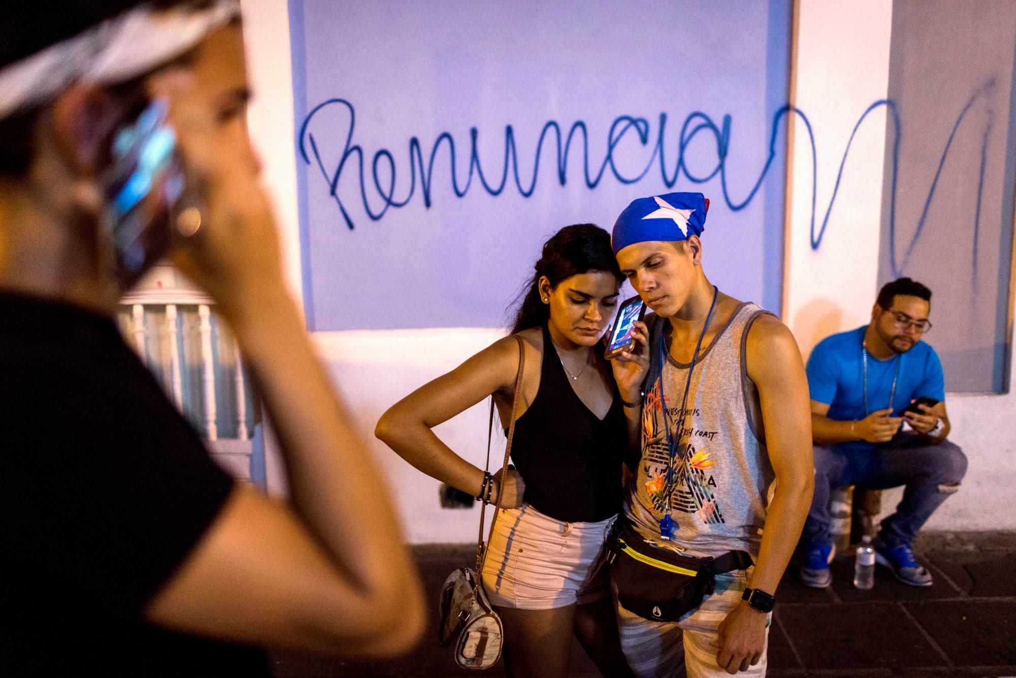 En esta imagen, tomada el 24 de julio de 2019, puertorriqueños escuchan en celulares un mensaje pregrabado del gobernador de Puerto Rico, Ricardo Rosselló, anunciando su renuncia el próximo 2  de agosto tras semanas de protestas luego de la filtracion de conversaciones de chat de contenido obsceno y misógino, en San Juan, Puerto Rico. 