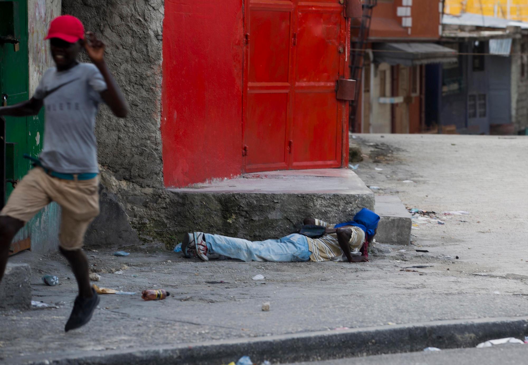 Un manifestante se acuesta en el suelo para cubrirse mientras otro huye al sonido de los disparos, durante una protesta para exigir la renuncia del presidente de Haití, Jovenel Moise, en el 216 aniversario de la Batalla de Vertieres en Puerto Príncipe, Haití, el lunes. 18 de noviembre de 2019. Al menos cuatro personas fueron baleadas y heridas durante una pequeña protesta en la capital de Haití después de un discurso del presidente Moise. Un periodista local, un oficial de policía y dos manifestantes fueron trasladados rápidamente con heridas de bala aparentes. 