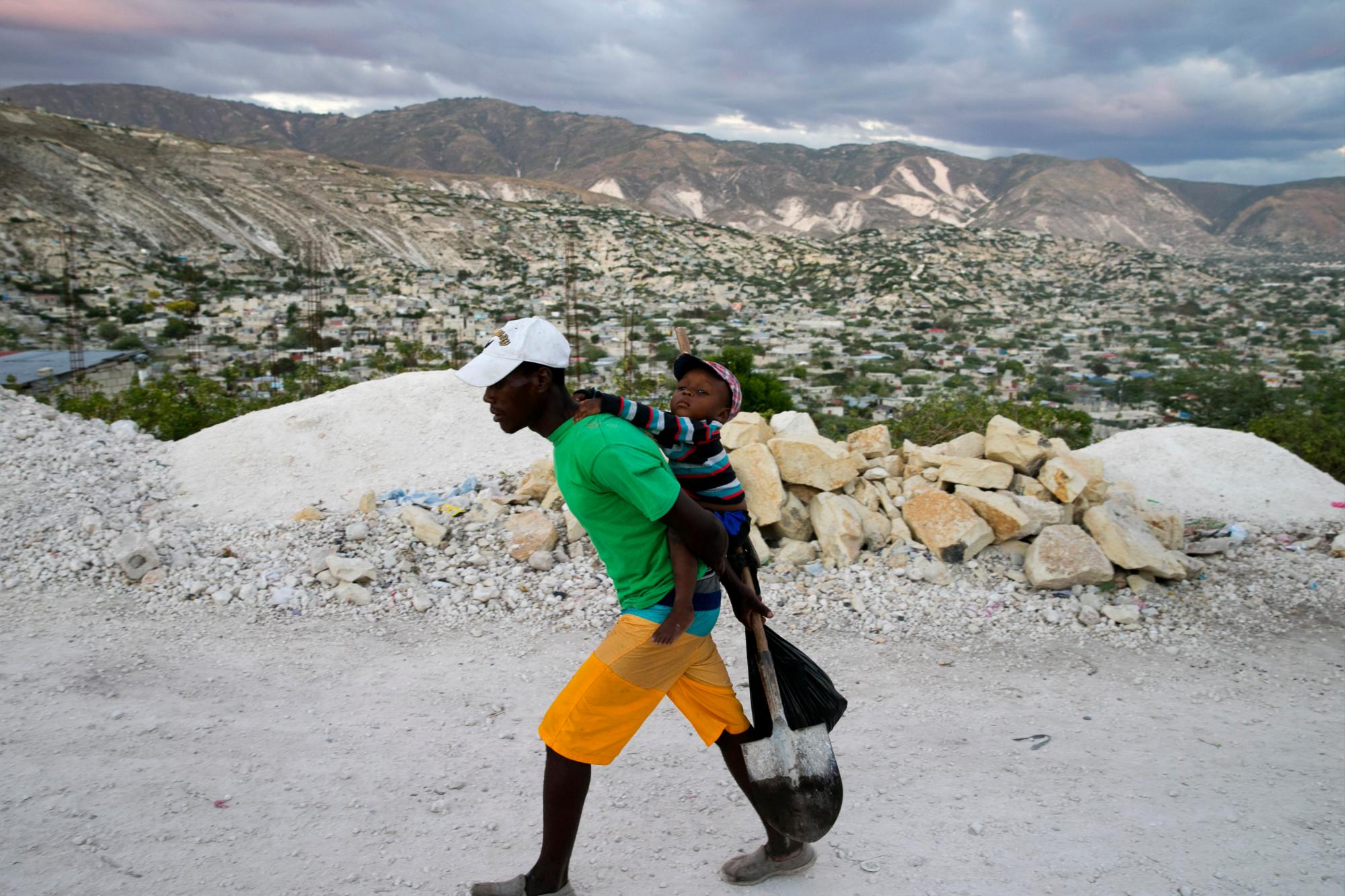En esta foto del 30 de diciembre de 2019, un trabajador de la construcción balancea a su hijo sobre su espalda mientras camina a casa con una pala después de trabajar donde vive en Canaan, un distrito en Croix des Bouquets, Haití, creado para las personas que perdieron sus hogares en El terremoto de hace 10 años. Ninguna de las administraciones recientes de Haití ha tomado medidas para mejorar las condiciones en Canaán, incluida la construcción de carreteras y la construcción de centros urbanos con servicios públicos que reducirían la dependencia de los habitantes de largos viajes a Puerto Príncipe, según Leslie Voltaire, un ciudadano urbano haitiano. planificador que ha trabajado para mejorar las condiciones en Canaan. 