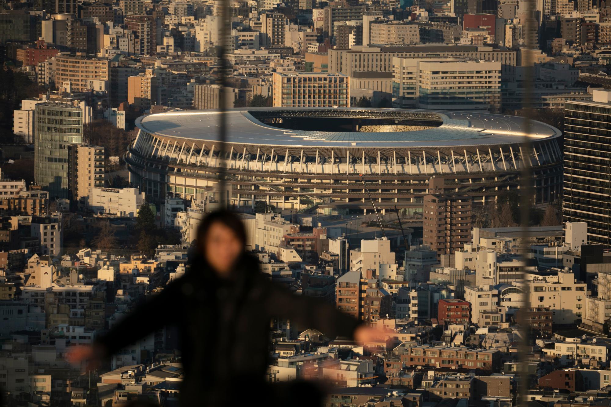 Un hombre en primer plano posa para fotos en la plataforma de observación Shibuya Sky mientras se ve el New National Stadium, un lugar para las ceremonias de apertura y clausura en los Juegos Olímpicos de Tokio 2020, el lunes 20 de enero de 2020, en el distrito de Shibuya de Tokio.