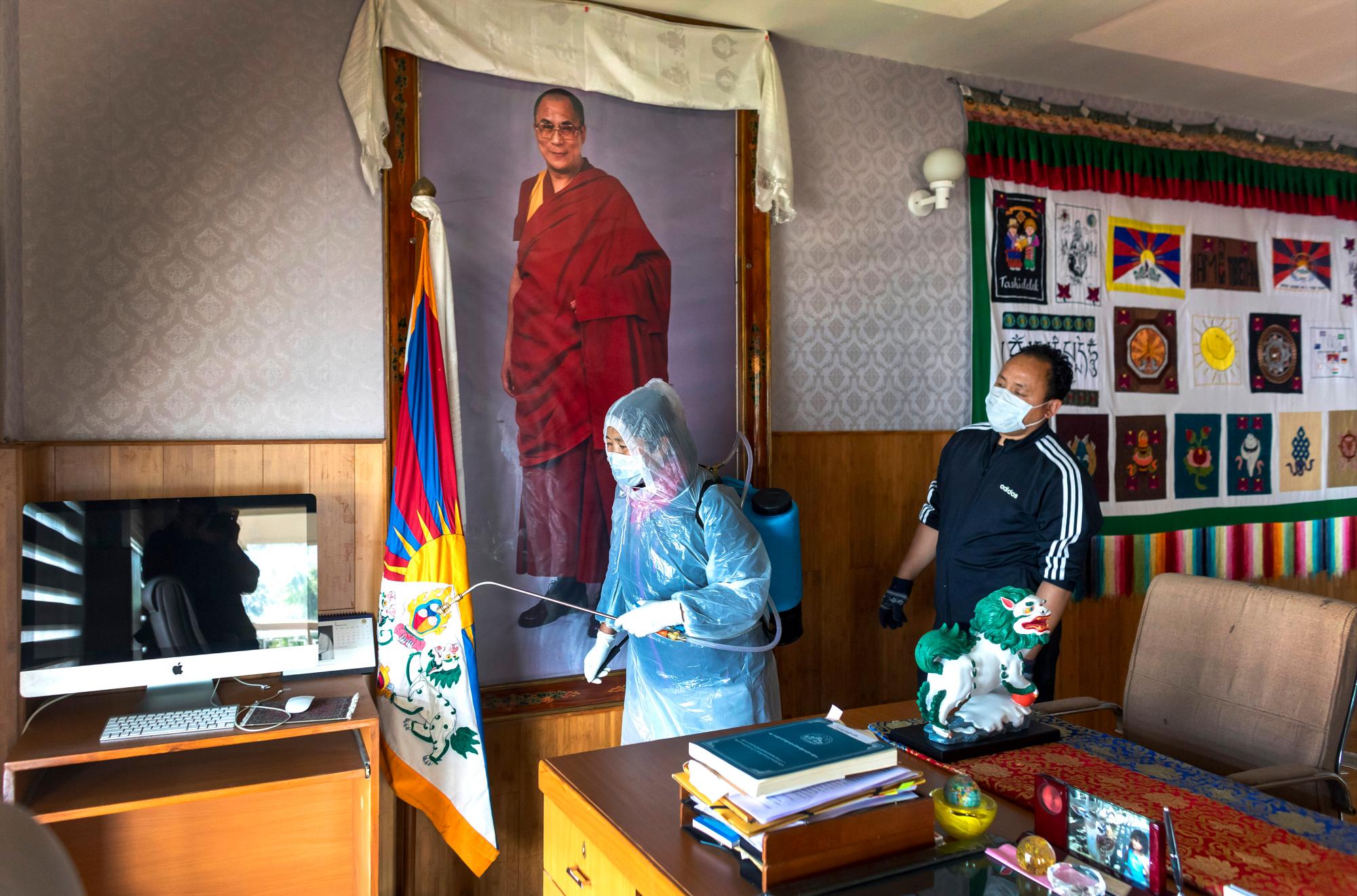 Un retrato del líder espiritual tibetano, el Dalai Lama, adorna una pared de la oficina del presidente tibetano Lobsang Sangay como empleado de la Administración Central Tibetana en ropa protectora rocía un esterilizador como precaución del coronavirus en Dharmsala, India, el jueves 19 de marzo. , 2020.