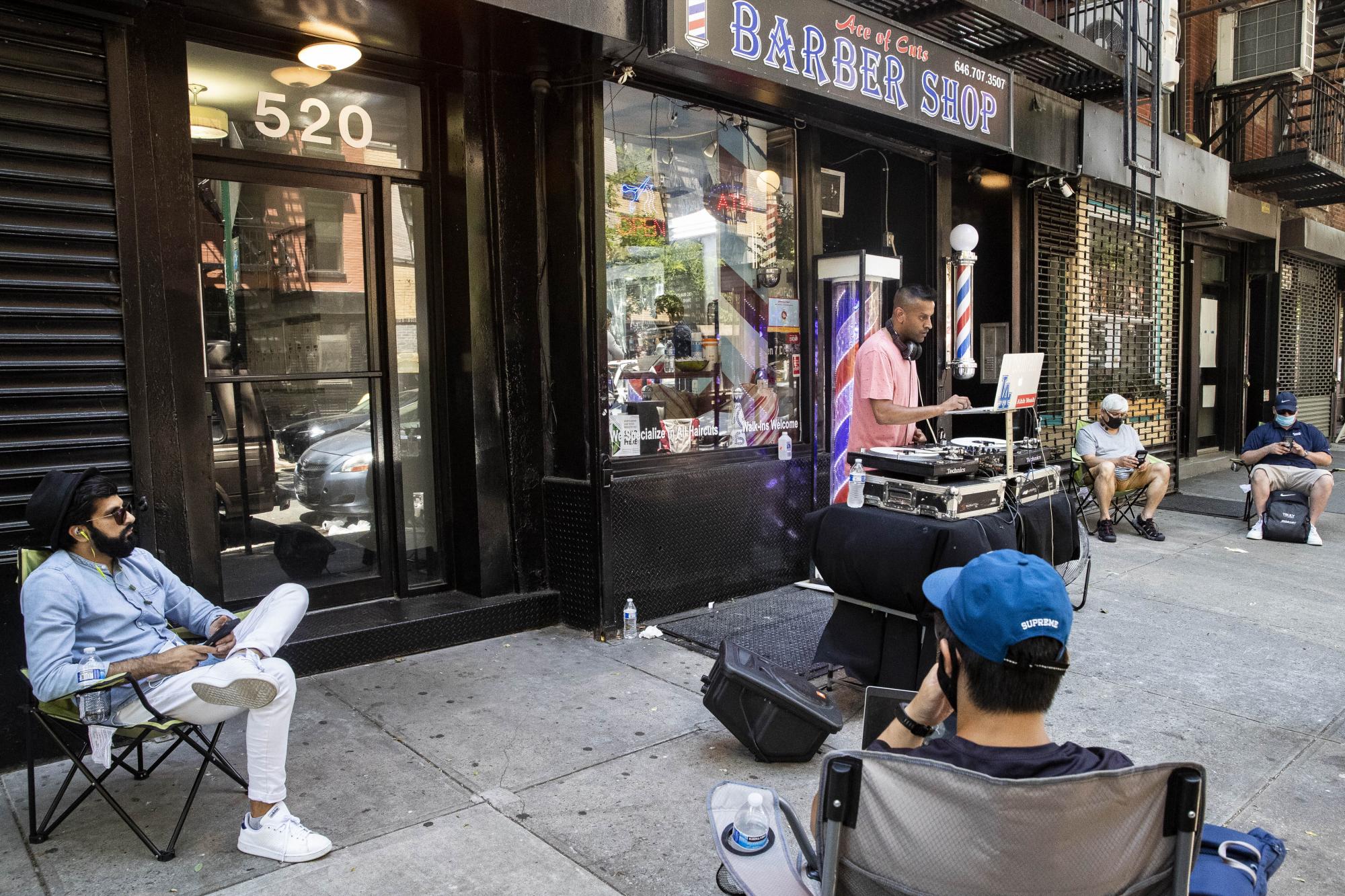 Cuatro clientes esperan en la acera frente a Ace of Cuts Barber Shop mientras un DJ ameniza la espera  (AP Photo/John Minchillo)