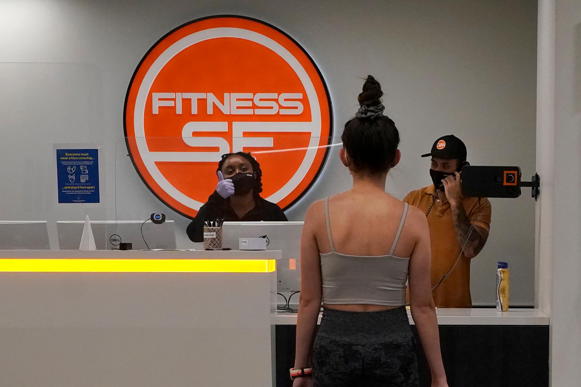 La trabajadora de recepción de Fitness SF Transbay, Taciana Fields, a la izquierda, levanta el pulgar mientras verifica la temperatura de un miembro del gimnasio que ingresa a las instalaciones durante el brote de coronavirus.
