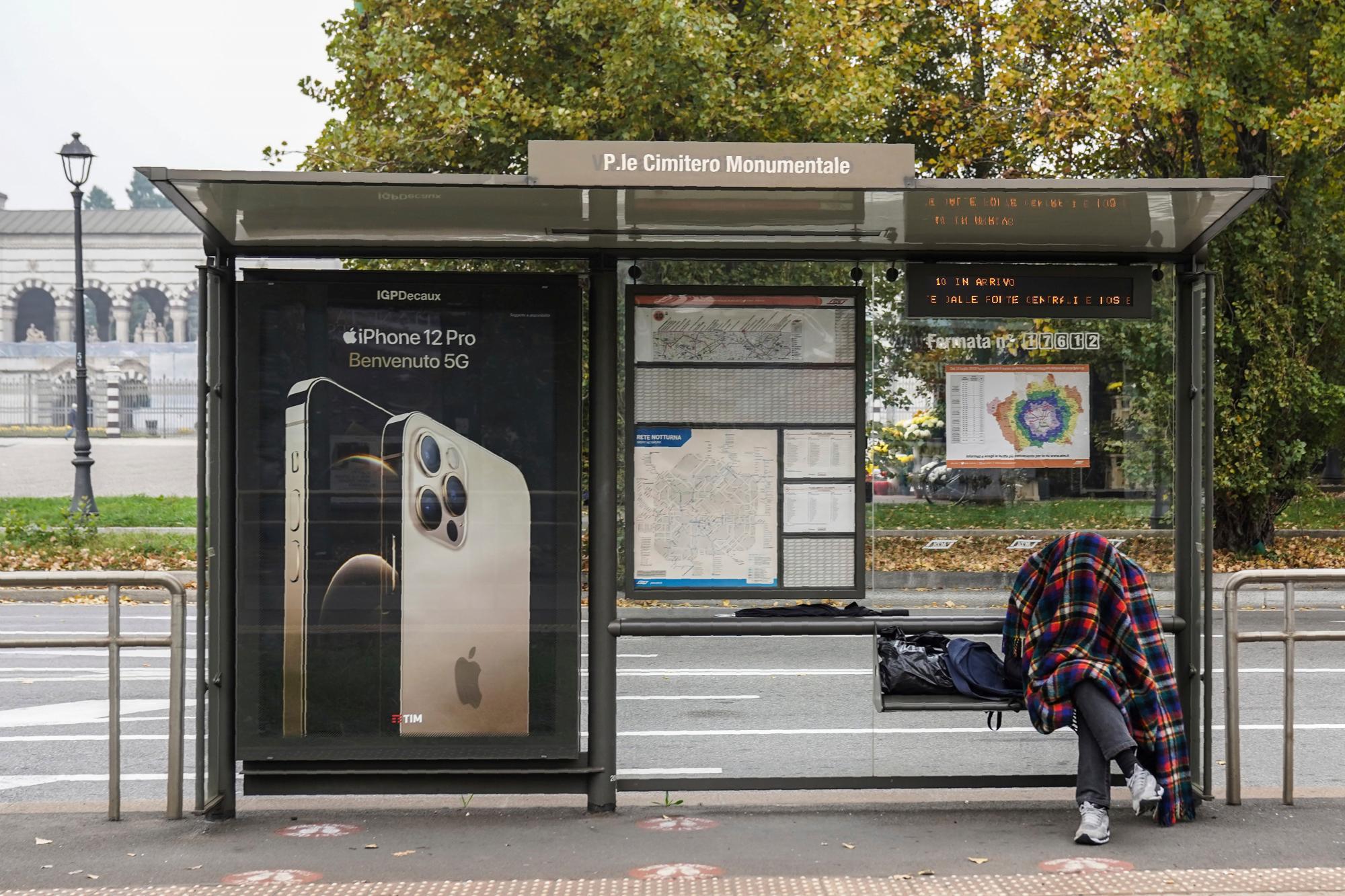 Un hombre se cubre con una manta mientras está sentado en un banco en una parada de tranvía durante un día gris y húmedo en Milán, Italia, el sábado 14 de noviembre de 2020 
