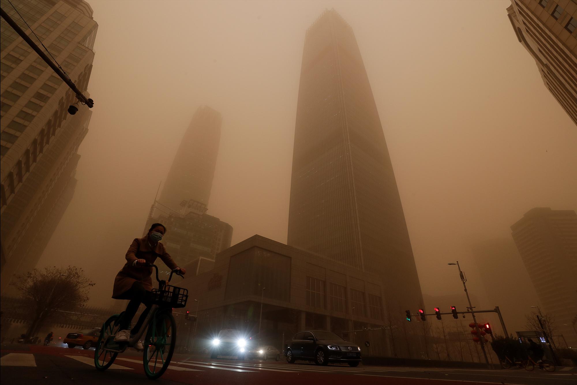 Un ciclista y automovilistas pasan por edificios de oficinas en medio de una tormenta de arena durante la hora pico de la mañana en el distrito comercial central de Beijing, el lunes 15 de marzo de 2021. La tormenta de arena trajo una neblina teñida a los cielos de Beijing y elevó los índices de calidad del aire el lunes. (AP Photo/Andy Wong)