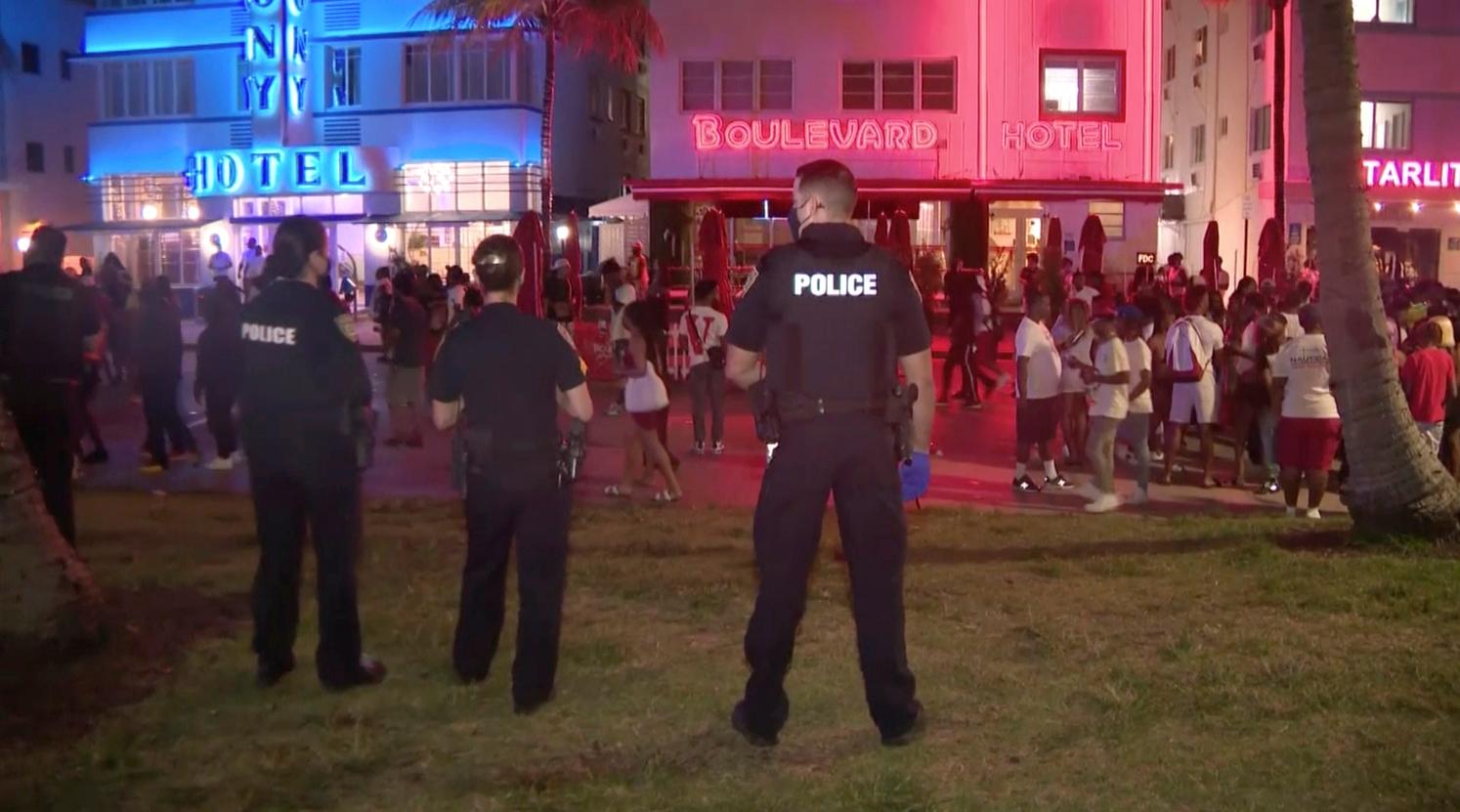 En esta imagen tomada del video, los oficiales de policía montan guardia el sábado 20 de marzo de 2021 por la noche, mientras la multitud desciende a South Beach en Miami. Después de días de fiesta y enfrentamientos entre la policía y grandes multitudes, los funcionarios de Miami Beach han ordenado un toque de queda de emergencia.