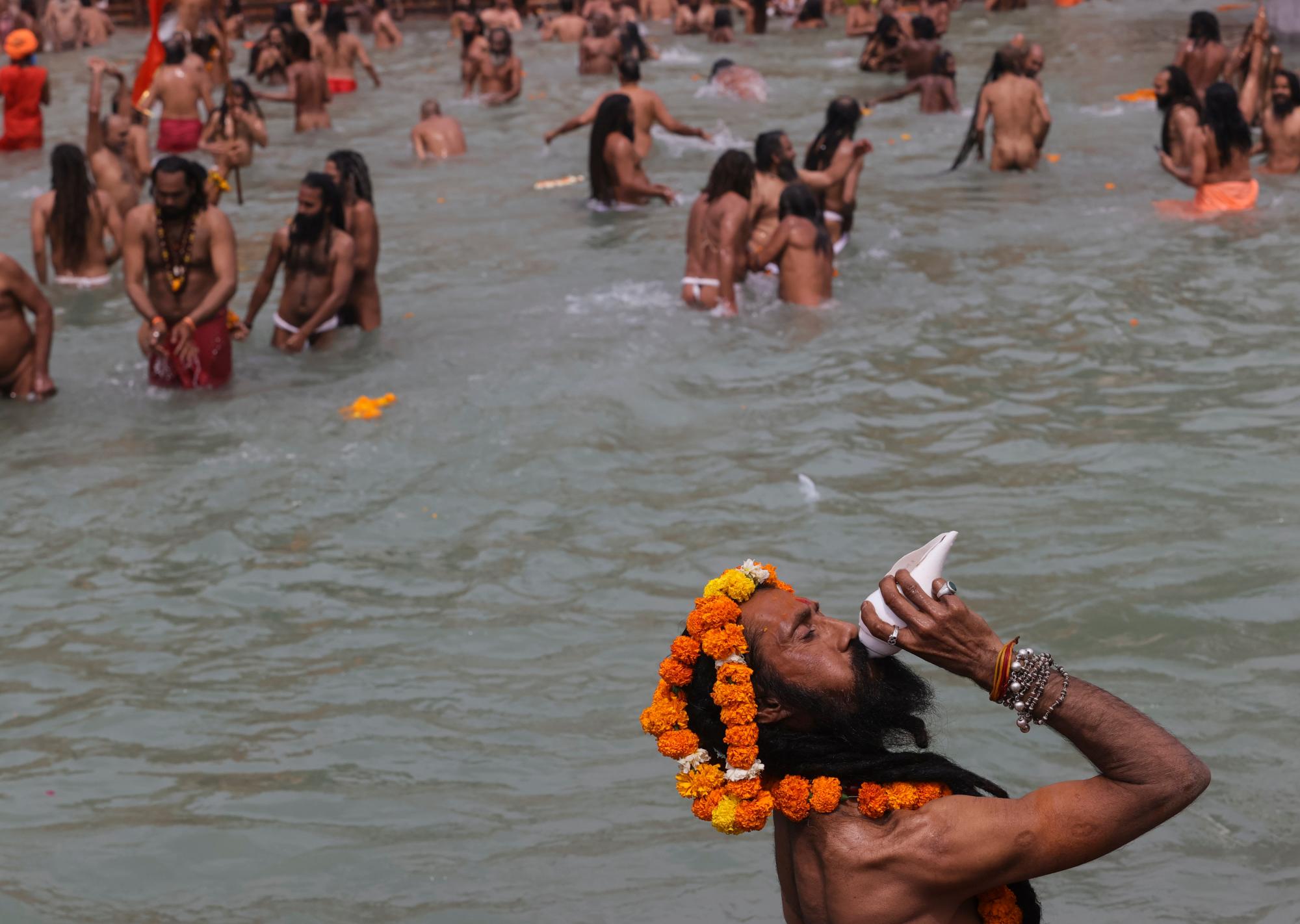 Un hombre santo hindú sopla una caracola mientras los devotos toman baños sagrados en el río Ganges durante el Kumbh Mela, o festival del cántaro, una de las peregrinaciones más sagradas del hinduismo, en Haridwar, estado norteño de Uttarakhand, India, el lunes 12 de abril de 2021 Decenas de miles de devotos hindúes se reunieron el lunes junto al río Ganges para oraciones especiales, muchos de ellos burlándose de las prácticas de distanciamiento social a medida que el coronavirus se propaga en la India a una velocidad récord. (AP Photo/Karma Sonam)