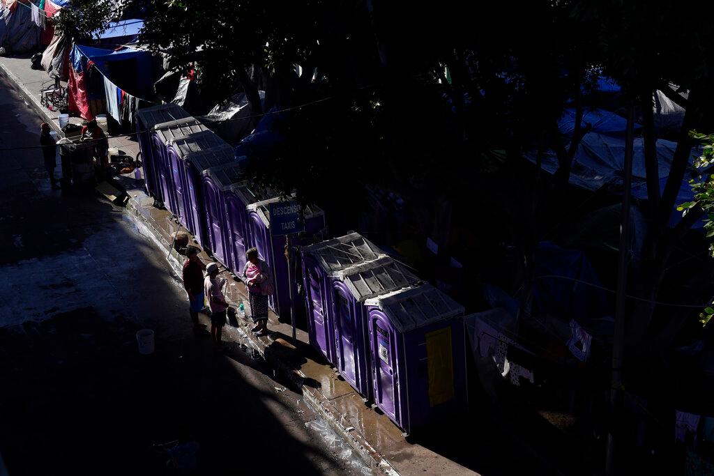 Migrantes charlan enfrente de los baños portátiles en un asentamiento que bloquea la entrada a un cruce peatonal a Estados Unidos (arriba) , el 8 de noviembre de 2021, en Tijuana, México. El campamento, ahora rodeado por una cerca metálica tras una operación policial el 28 de octubre, sirve de alojamiento temporal a cientos de migrantes que esperan solicitar asilo en Estados Unidos.