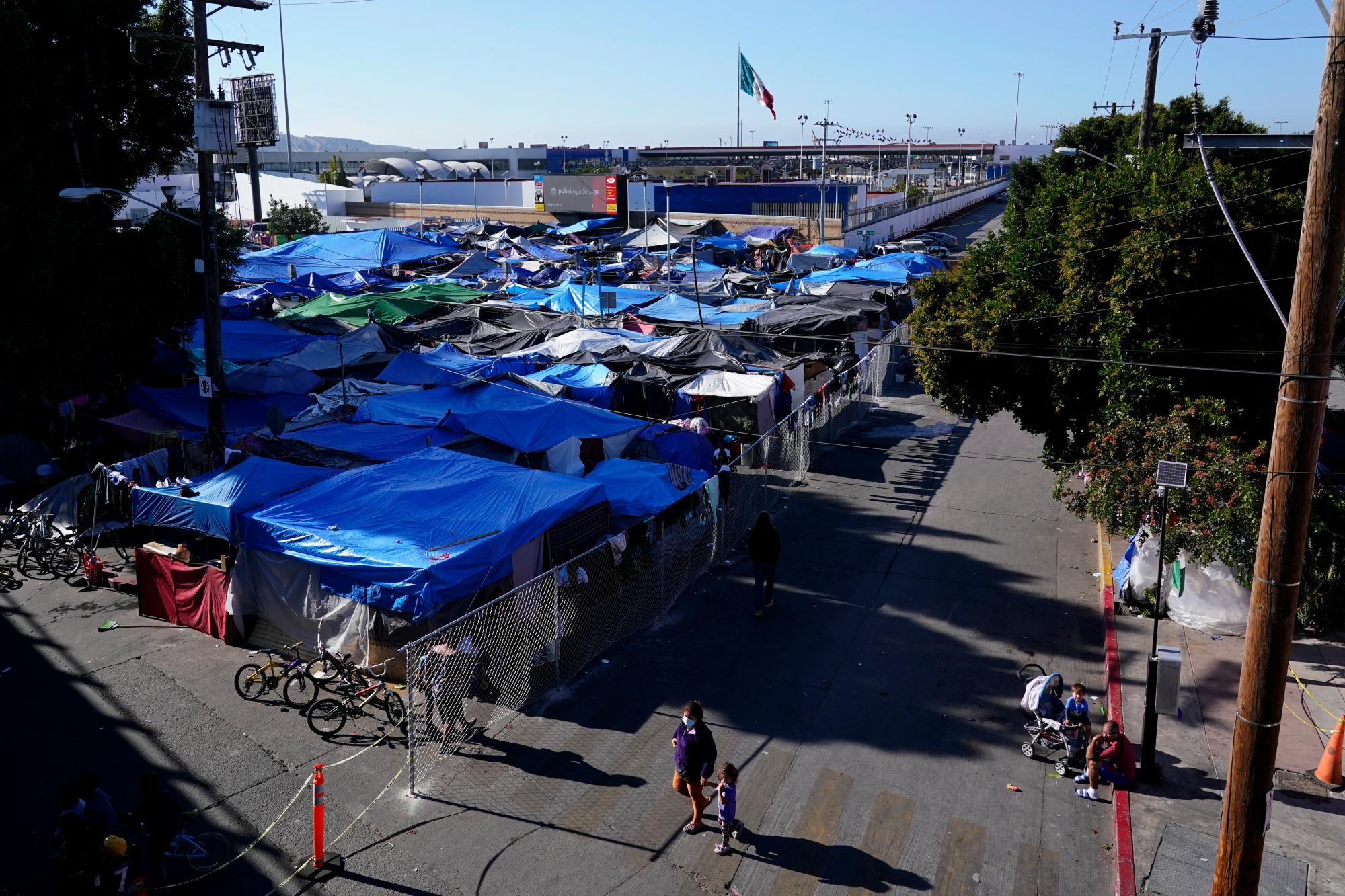 Migrantes (abajo) caminan por las inmediaciones de un asentamiento que bloquea la entrada a un cruce peatonal a Estados Unidos (arriba), el 8 de noviembre de 2021, en Tijuana, México. El campamento, ahora rodeado por una cerca metálica tras una operación policial el 28 de octubre, sirve de alojamiento temporal a cientos de migrantes que esperan solicitar asilo en Estados Unidos.