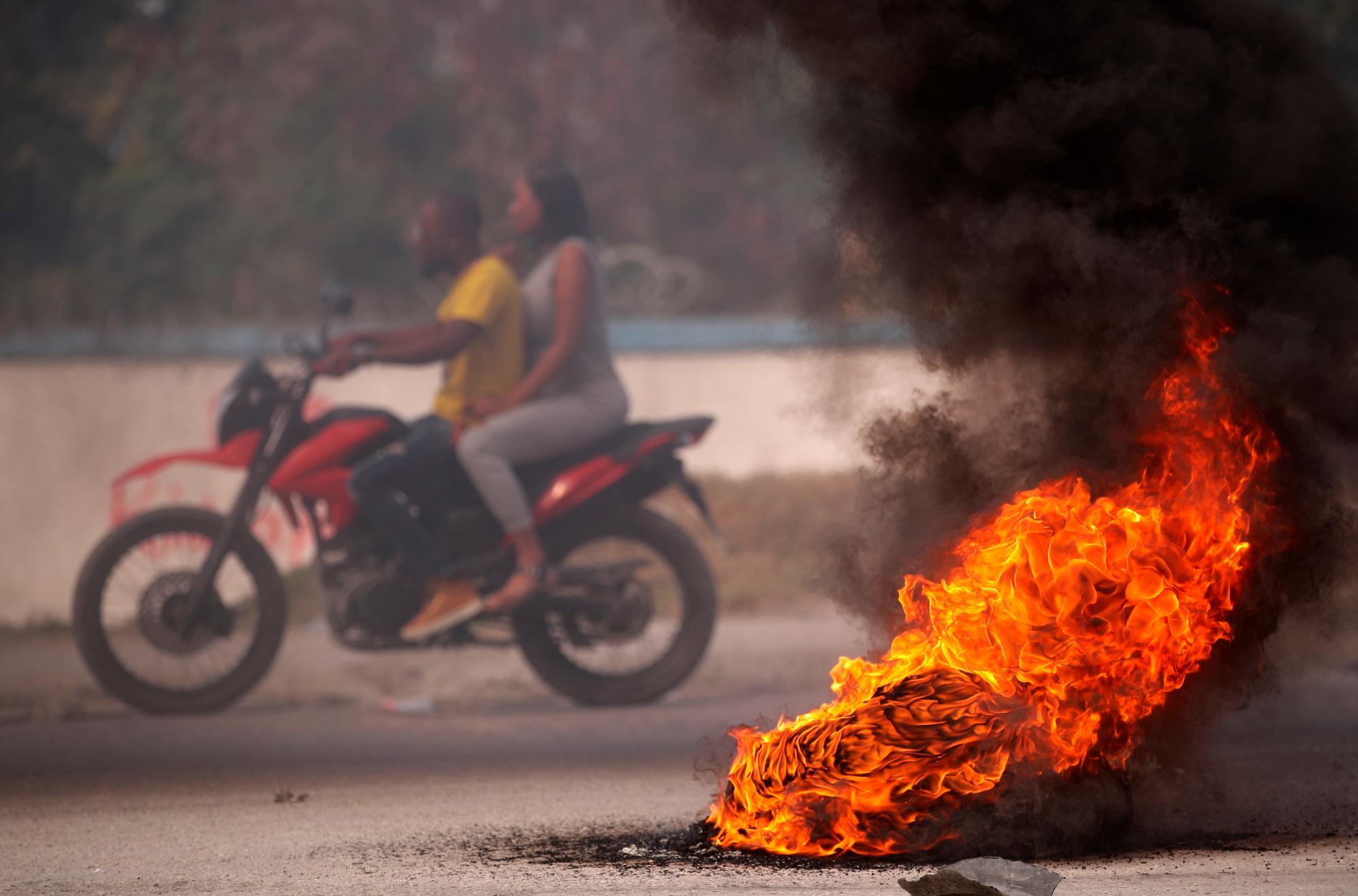 PUERTO PRÍNCIPE (HAITÍ), 11/02/2021.- Personas cruzan hoy en motocicleta cerca a una barricada en llamas, durante una protesta contra el Gobierno en Puerto Príncipe (Haití). Haití sufre por estos días una nueva crisis política tras la jubilación forzosa de tres jueces opositores ordenada el lunes por el Gobierno de Jovenel Moise, en aparente violación de la Constitución. EFE/ Orlando Barría