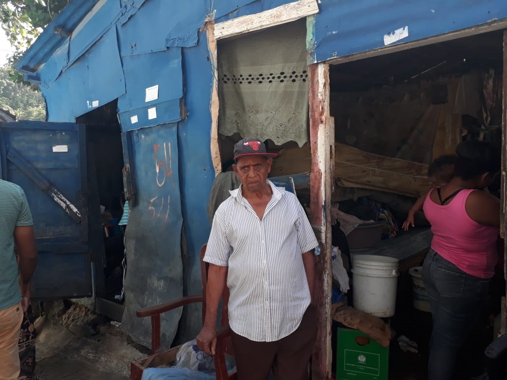 El señor Santos Méndez frente a su vivienda y una nieta adolescente que a su vez tiene un bebé, mientras esperan que le hagan la mudanza hacia Las Américas.