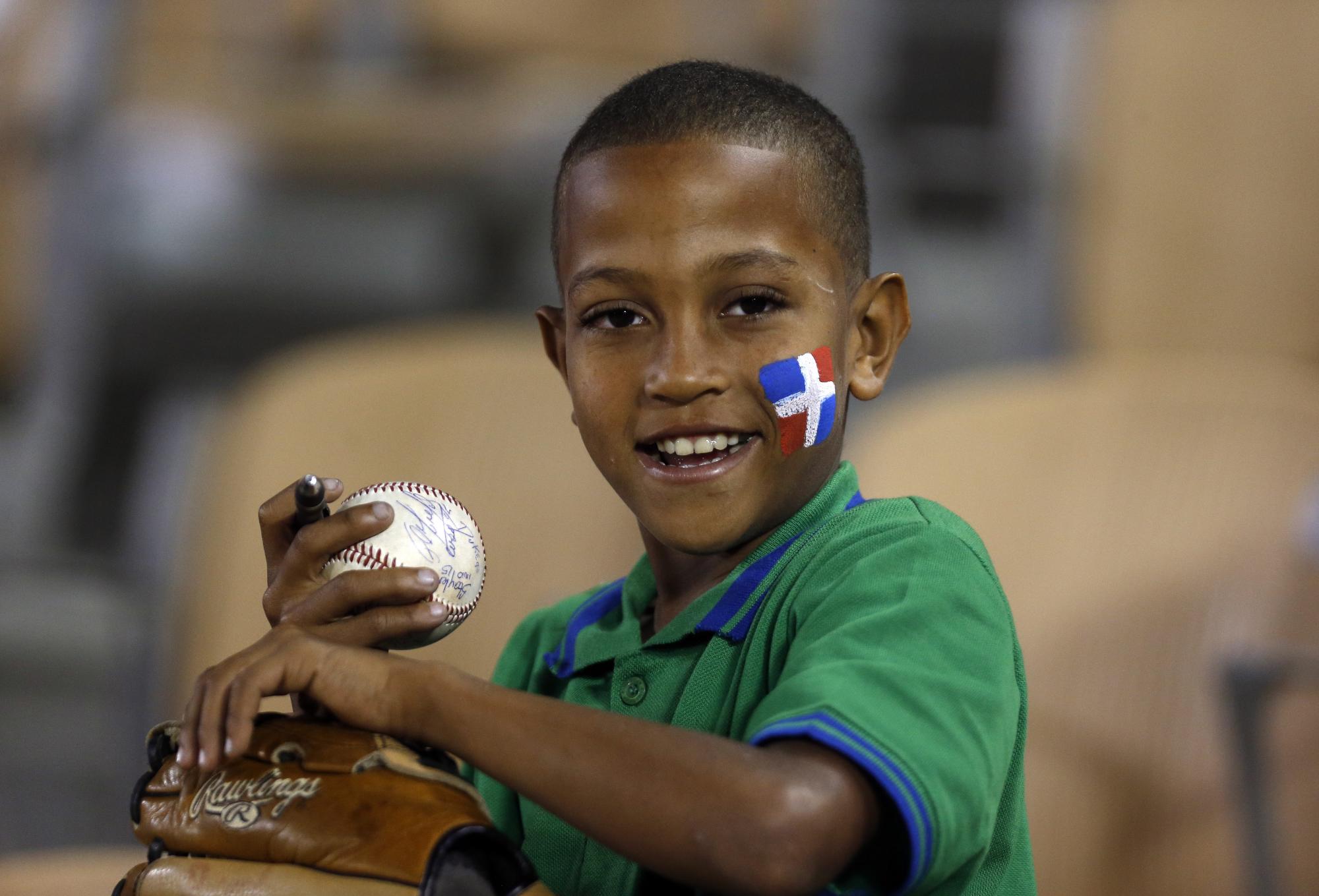 El fanático o pelotero del futuro, un niño, que sostiene bola autografiada, su guante y la expresión de la dominicanidad con su bandera en la mejilla, acompañada de una inocente sonrisa.