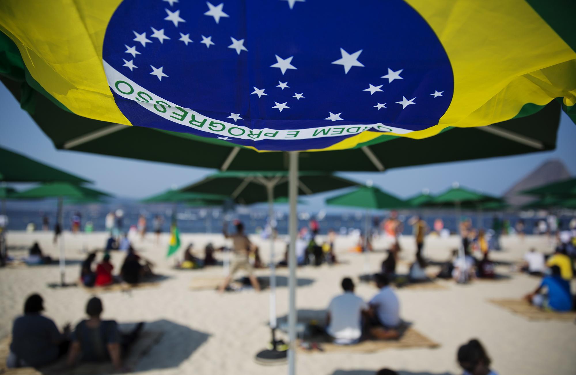 La bandera de Brasil ondea bajo una sombrilla mientras los espectadores observan desde una playa durante las competiciones de vela.